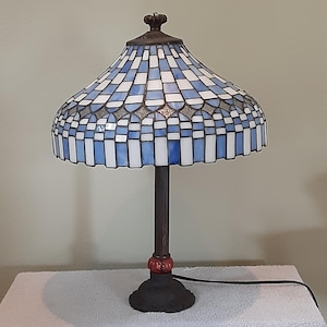 May include: A Tiffany-style table lamp with a blue and white stained glass shade. The shade has a geometric pattern and is supported by a dark brown metal base with a decorative red accent. The lamp is plugged into an electrical outlet.
