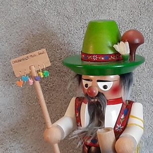 May include: A wooden nutcracker figurine in the form of a man, wearing a green hat with a red and white floral band, a white shirt, and red suspenders with floral designs. The figurine holds a wooden staff with heart-shaped charms and a beer stein.