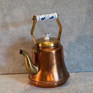May include: A copper tea kettle with a brass spout and handle supports. The handle is white with blue floral designs. The lid is brass with a white knob. The kettle has a vintage aesthetic.