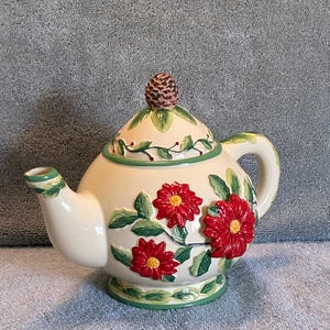 May include: Cream-colored ceramic teapot with red flower and green leaf accents. The lid features a pinecone-shaped handle. The teapot has a curved spout and handle, and sits on a green-trimmed base. A decorative piece for tea lovers.