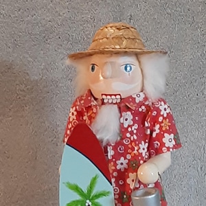 May include: A wooden nutcracker figure wearing a straw hat and a red short-sleeved shirt with a white floral pattern. The figure has white hair and a beard, holds a light blue surfboard with a green palm tree graphic, and carries a small silver bucket.