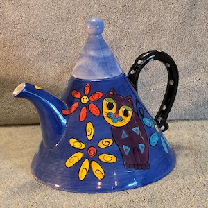 May include: A blue ceramic teapot with a whimsical cat design. The teapot features a black handle with white polka dots, a blue lid, and is decorated with colorful flowers and a cat illustration.