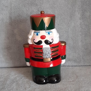 May include: A ceramic Christmas nutcracker figurine. The figurine is red, green, and gold with a white beard and mustache. It has blue eyes and a black hat. The nutcracker is a festive holiday decoration.