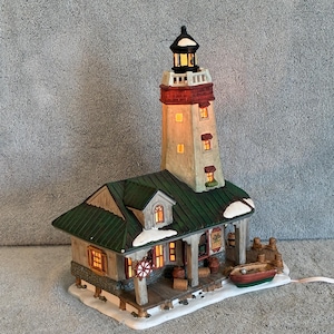 May include: A detailed miniature lighthouse with a connected building. The lighthouse has a red top and a black lantern. The building has a green roof, a wooden porch, and a small boat. The scene is illuminated with warm, inviting lights.