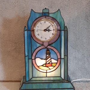 May include: A stained glass table clock featuring a lighthouse design. The clock face is round with Roman numerals. The lighthouse is depicted in a circular window, with a blue and white sky. The clock is approximately 10 inches tall.