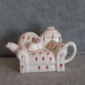 May include: White ceramic teapot shaped like a couch with a white cat sitting on it. The couch has a pink and green floral pattern.