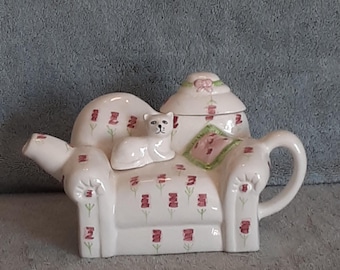 May include: White ceramic teapot shaped like a couch with a white cat sitting on it. The couch has a pink and green floral pattern.