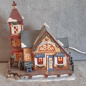 May include: A miniature ceramic Seaside Cafe with a light-up interior. The building features a brown exterior, a gray roof, and a red tower. The sign reads "Seaside Cafe" and a small chalkboard advertises "Special Lobster Dinner".