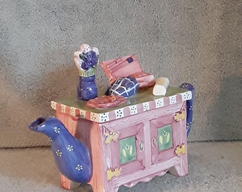 May include: A whimsical teapot shaped like a pink and purple cabinet with a blue spout and handle. The teapot has a green top with white stripes and is decorated with floral accents. A small vase with flowers and other decorative items sit on top.