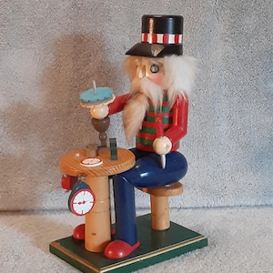 May include: A wooden nutcracker figurine depicting a craftsman at work. The figure wears a red jacket with green stripes, blue pants, and a black hat with red and white stripes. The figurine is holding tools and is seated at a small wooden table with a clock.