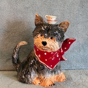 May include: A ceramic Yorkshire Terrier figurine with a red polka-dot bandana and a tiny teacup on its head. The dog is primarily gray and brown with a cream-colored face. The teacup is white with red polka dots and a blue floral design.