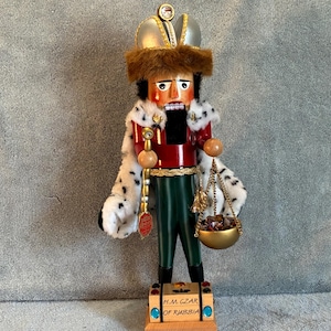 May include: A nutcracker figure dressed as a czar, wearing a silver crown with a faux fur trim, a red jacket, and a white faux fur cape with black spots. The figure holds a gold staff and a gold scale with small stones, standing on a base labeled H.M. CZAR OF RUSSIA.