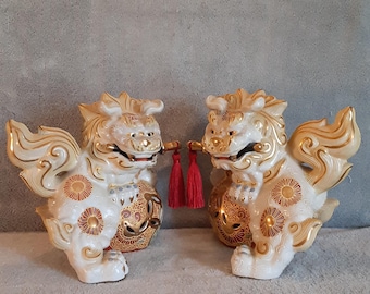 May include: Pair of white ceramic Foo Dogs with gold accents. Each dog holds a ball and has gold detailing on the mane, wings, and body. Red tassels hang from their mouths. Decorative figurines.