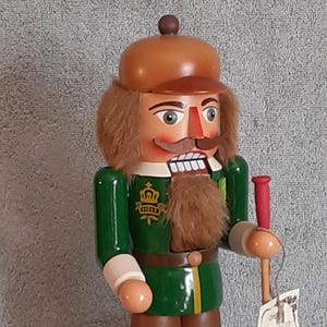 May include: A wooden nutcracker figurine in a green and brown uniform with a brown cap and faux fur hair and beard. The figurine holds a red and wooden staff. A gold crown emblem is on the chest. The nutcracker has a detailed painted face.