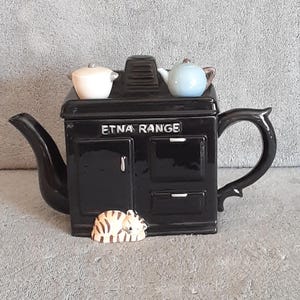 May include: Black ceramic teapot shaped like a stove, with the text "ETNA RANGE" on the front. It has a small white and blue teapot on top, and a ceramic cat figurine in front.