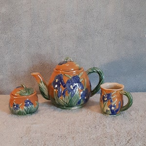 May include: A brown teapot, sugar bowl, and creamer set with a floral design of blue irises and green leaves.