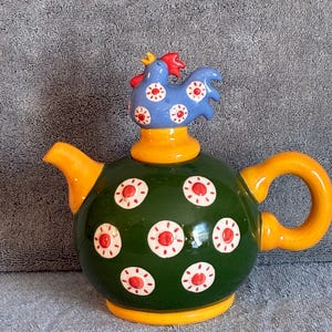 May include: A round ceramic teapot with a dark green body featuring white and red circular floral patterns. The teapot has a bright yellow handle, spout, and base. The lid is topped with a blue rooster figure with a red comb and white floral accents.
