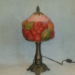 May include: A table lamp with a frosted glass shade featuring a relief design of red grapes and green leaves. The shade has a pink top section and a pointed finial. The base is made of dark bronze-colored metal with an ornate, flared design.