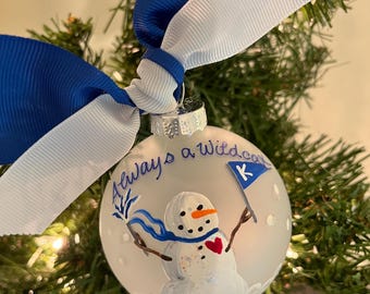 May include: A white glass Christmas ornament with a painted snowman holding a blue and white flag with a 'K' on it. The ornament is decorated with white glitter and blue snowflakes. The ornament is tied with a blue and white ribbon and reads "Always a Wildcat".