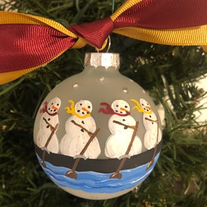 May include: A gray glass Christmas ornament with a hand-painted design of four snowmen rowing a boat on a blue lake. The snowmen are wearing scarves and hats, and the boat is black with white oars.