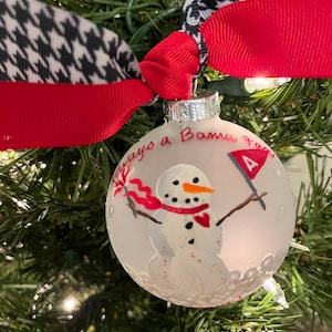 May include: White glass Christmas ornament with a snowman painted on it. The snowman is wearing a red scarf and holding a red and white flag with the letter "A" on it. The ornament has the words "Always a Bama Fan" written on it.
