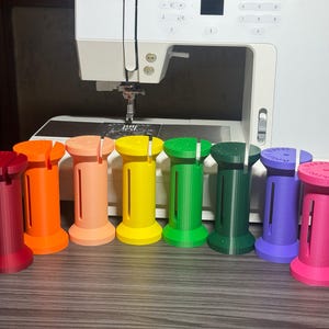 May include: A set of eight cylindrical plastic thread spools in a rainbow of colors including red, orange, peach, yellow, green, dark green, purple, and pink. Each spool features a vertical slit and a flat top with printed text.
