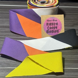 May include: A roll of fabric binding featuring a geometric pattern of purple, orange, white, black, and yellow segments. A square brown tag is attached, displaying a pink circular sticker with the text Meise's Pieces RIBBON CANDY BINDING.