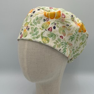 May include: A patterned scrub cap featuring a botanical design with gourds, leaves, and berries in shades of yellow, green, and purple on a cream background. The cap is made of fabric and is designed to be worn on the head.