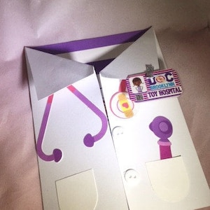 May include: A white paper doctor's coat with purple accents, including a stethoscope design and a pocket. The coat features a "Brooklyn Toy Hospital" badge and a bandage sticker. The coat is folded and appears to be a greeting card.