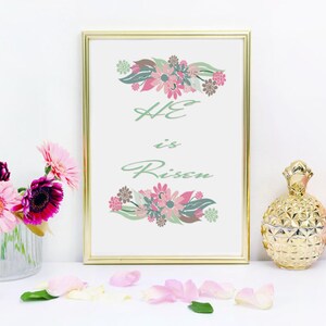 May include: A gold framed print with a white background and green and pink floral designs. The text "HE is Risen" is written in green cursive lettering. The print is sitting on a white surface with pink flowers, a gold pineapple shaped jar, and a pink candle.