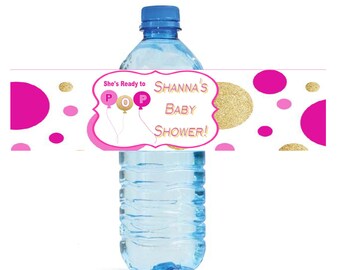May include: A clear plastic water bottle with a blue cap and a decorative label. The label features the text "Shanna's Baby Shower!" with pink and gold accents, including polka dots and balloon graphics. The bottle is filled with clear liquid.