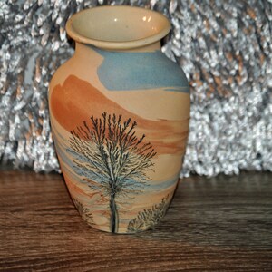 May include: A small ceramic vase with a light brown, blue, and orange swirl design. The vase features a black silhouette of a tree with branches.
