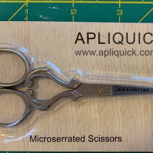 May include: A silver pair of microserrated scissors with decorative handles on a brown and white background. The packaging includes the brand name "APLIQUICK® TM" and the website "www.apliquick.com".