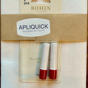 May include: A package of two Apliquick tools by Bohin France, featuring white and red cylindrical applicators on a branded card. The set includes a large white sheet of stabilizer material, with text reading APLIQUICK, 98477, 2 pcs, and Kathy McNeil ART QUILTS.