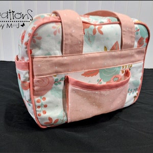 Pode incluir: Uma bolsa retangular de tecido com fundo branco e estampa floral em tons de pêssego, azul-petróleo e verde. A bolsa possui alças, acabamentos e um bolso frontal na cor pêssego sólido. O texto cReations by M and J é visível.