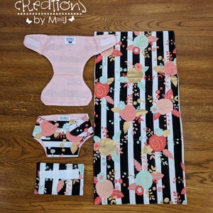 May include: A set of baby cloth diapering items. Includes a pink diaper with white polka dots, a diaper cover, a changing pad, and a wipes case. The diaper cover, changing pad, and wipes case feature a black and white striped pattern with floral accents.