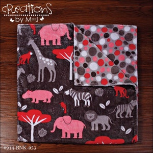 May include: A reversible baby blanket with a brown background featuring a jungle animal print on one side and a gray and pink polka dot pattern on the other side. The animal print side features pink elephants, giraffes, zebras, hippos, lions, and monkeys. The text "Creations by Mandi" is printed in the top left corner.