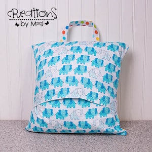 Pode incluir: Uma bolsa de armazenamento de tecido azul e branco com uma al&ccedil;a colorida. A bolsa apresenta um padr&atilde;o repetido de elefantes azuis com bolinhas brancas. A bolsa &eacute; etiquetada como "Creations by M and J".