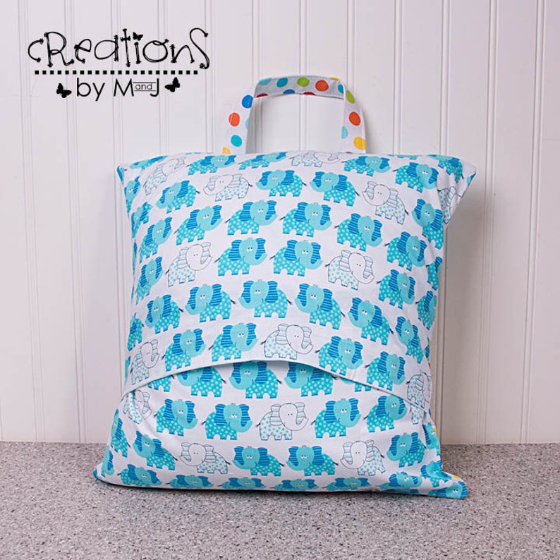 Pode incluir: Uma bolsa de armazenamento de tecido azul e branco com uma al&ccedil;a colorida. A bolsa apresenta um padr&atilde;o repetido de elefantes azuis com bolinhas brancas. A bolsa &eacute; etiquetada como "Creations by M and J".