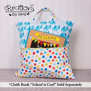 Pode incluir: Uma bolsa de tecido branca para livros com uma estampa de elefantes azul e branco na frente. A bolsa tem um bolso na frente com um pequeno livro dentro. O livro tem um fundo marrom com a palavra "WELCOME" em letras amarelas. A bolsa tem um padr&atilde;o de bolinhas na parte inferior.