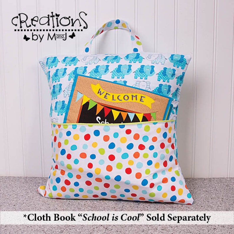 Pode incluir: Uma bolsa de tecido branca para livros com uma estampa de elefantes azul e branco na frente. A bolsa tem um bolso na frente com um pequeno livro dentro. O livro tem um fundo marrom com a palavra "WELCOME" em letras amarelas. A bolsa tem um padr&atilde;o de bolinhas na parte inferior.