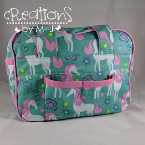 Pode incluir: Uma bolsa de tecido verde-azulado com um padrão repetitivo de unicórnios brancos com crinas e caudas cor-de-rosa, acentuado por estrelas, corações e flores. A bolsa inclui debrum cor-de-rosa, duas alças superiores e um bolso frontal com borda rosa.