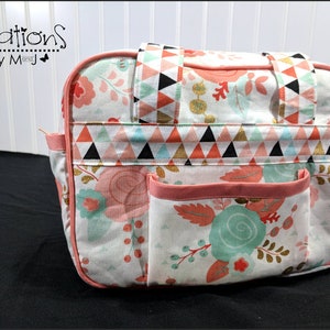 Pode incluir: Uma bolsa de tecido retangular com base branca e padrão floral em tons de coral, verde menta e dourado. A bolsa inclui uma faixa horizontal com estampa geométrica de triângulos, duas alças superiores e um bolso frontal com debrum coral.
