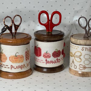 Puede incluir: Tres contenedores de madera con cubiertas de tela bordada y tijeras de diferentes colores en la parte superior. Los contenedores están etiquetados como "Pumpkin", "Tomato" y "Snip Snip".