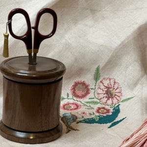 以下が含まれることがあります： 木製の糸ホルダーと、バーガンディ色のハサミ。 ハサミは金色の刃と小さな金のタッセルが付いています。 糸ホルダーは円筒形で、上部と下部が丸みを帯びています。 背景には刺繍の花があります。