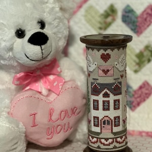 May include: A decorative wooden spool featuring a cross-stitched house design with red and gray accents. Two white doves flank a heart and envelope. A white teddy bear with a pink bow and a pink heart that says "I love you" is in the background.