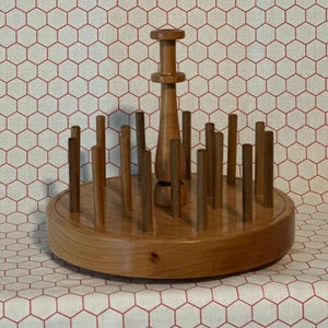 May include: A circular wooden stand with a smooth, polished finish featuring a central vertical post and numerous smaller upright pegs arranged in a ring. The base is thick and cylindrical, providing a stable foundation for the upright wooden pegs.