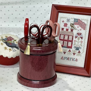 May include: A wooden scissor holder with a red, white, and blue theme. The holder has a red, white, and blue tassel and is filled with four pairs of scissors. The holder is sitting on a white surface with a red and white cross-stitch pattern. The cross-stitch pattern is framed in a red wooden frame. The cross-stitch pattern features a house with a red roof and a flag with the words "America" underneath.