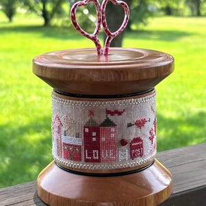 May include: A wooden thread spool with a cross-stitch design featuring red houses, a mailbox, and the word "LOVE". The spool is topped with two red and white heart-shaped wire ornaments.