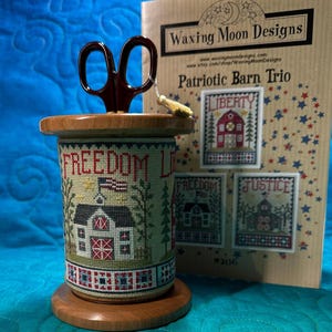 May include: A wooden thread holder with a cross-stitch design featuring a barn, American flag, and the words "FREEDOM LIBERTY". A pair of brown scissors sits atop the holder. A booklet titled "Patriotic Barn Trio" is in the background.